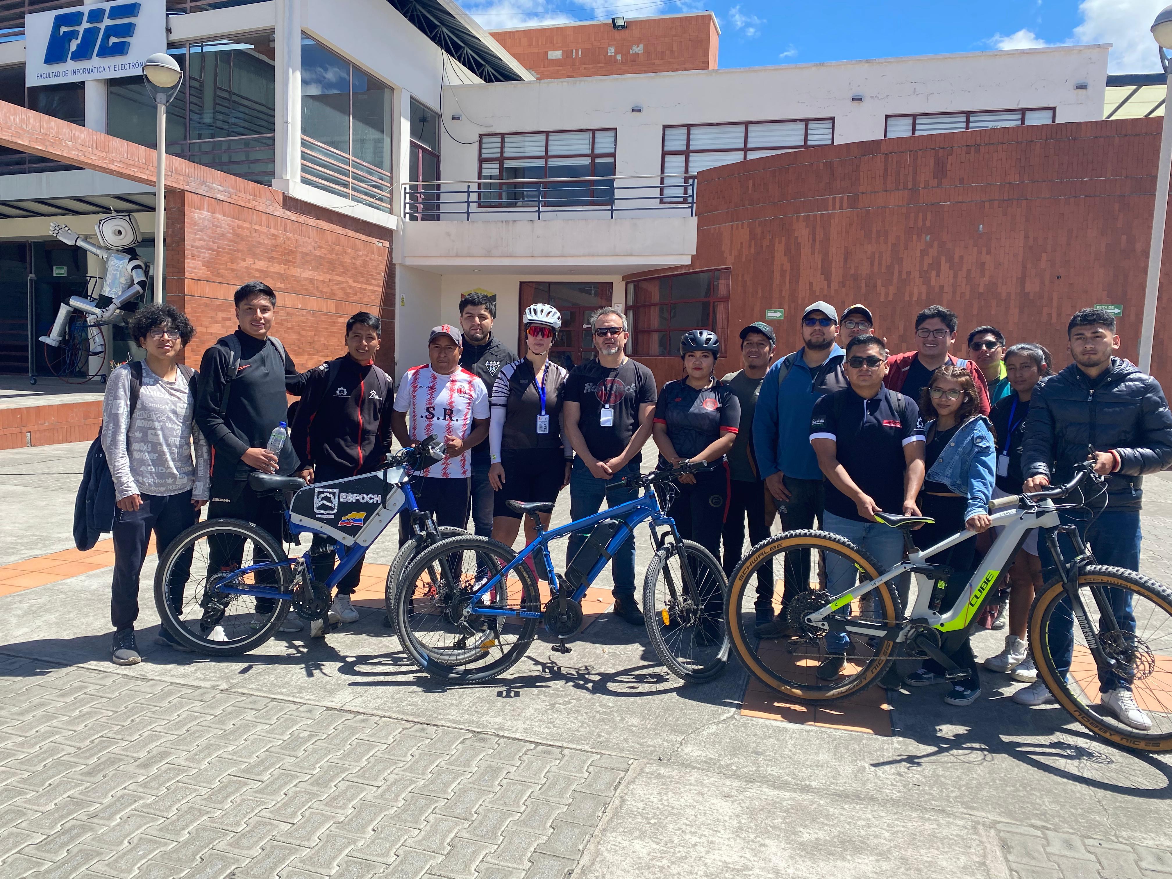 Group photo of cyclists smiling with bicycles in front of a brick building on a sunny day.